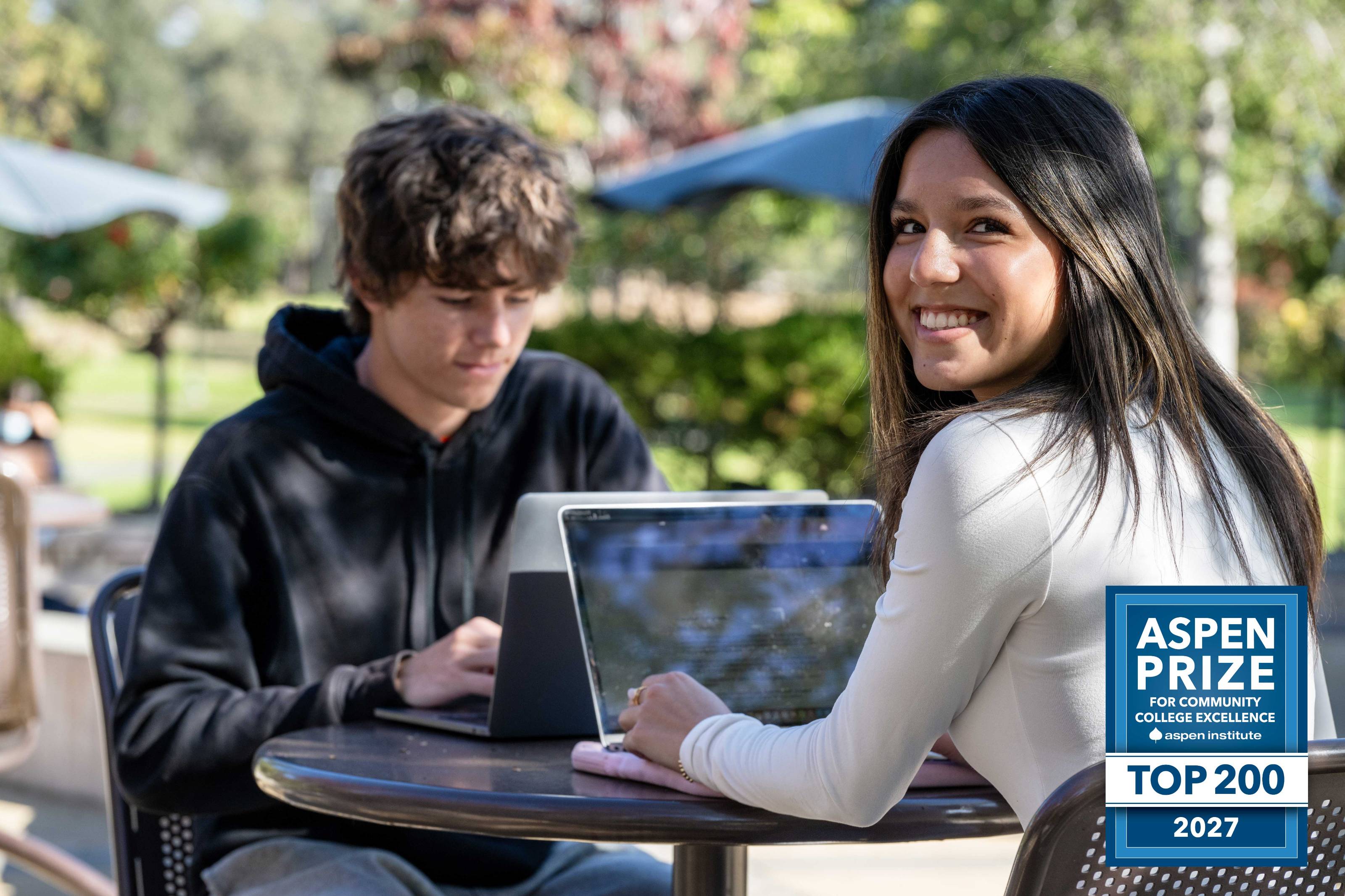 Two people sitting at an outdoor table on a sunny day, each using a laptop. The setting includes green trees, umbrellas, and blurred foliage in the background. A blue badge in the lower right corner reads: “Aspen Prize for Community College Excellence – Top 200 – 2027.