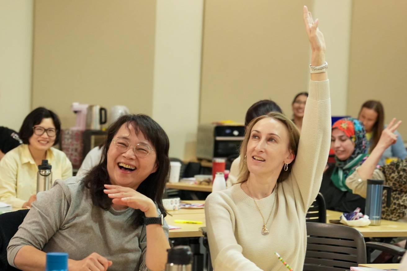 left- esl student with dark brown hair and glasses laughing. right- esl student with blonde hair raising her hand