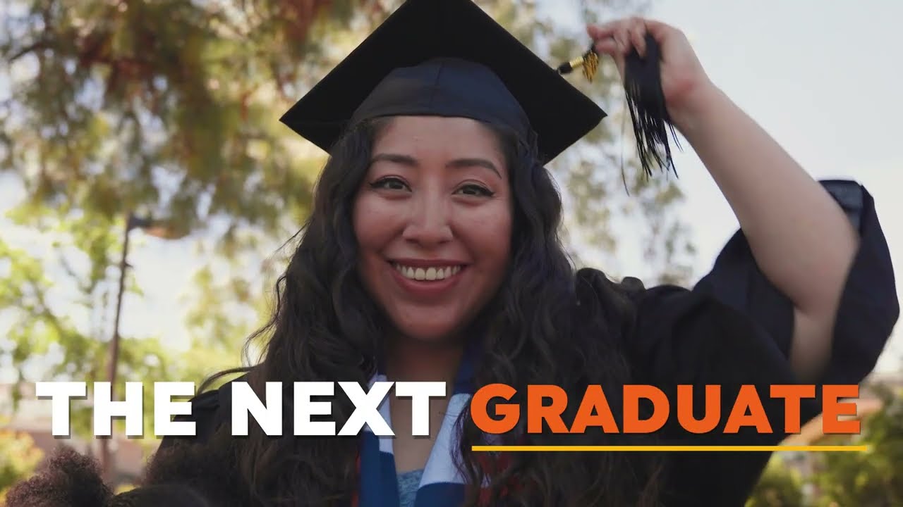 female student with long brown hair wearing a cap and gown holding her tassel