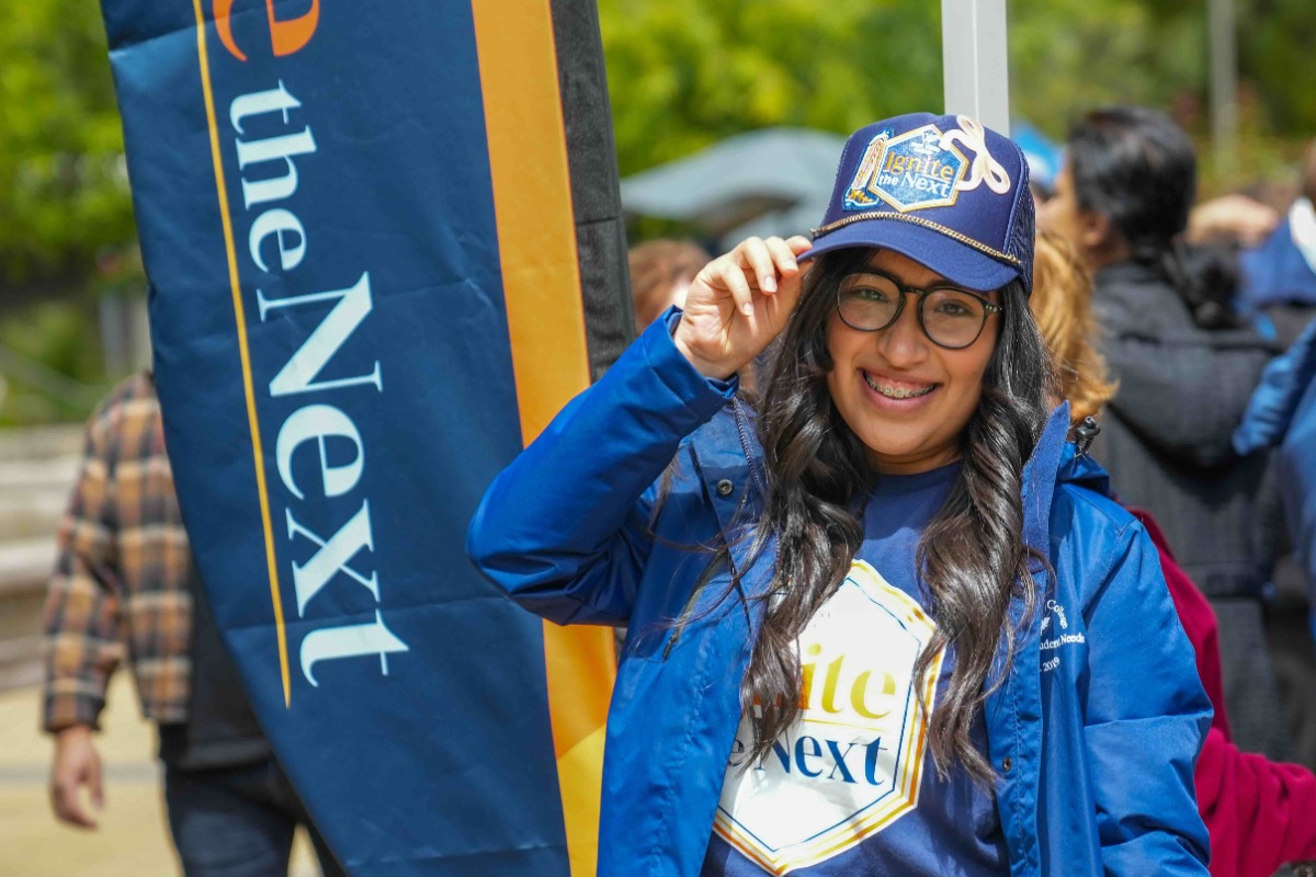 female student with long brown hair wearing black glasses wearing a blue ignite the next branded hat and tshirt